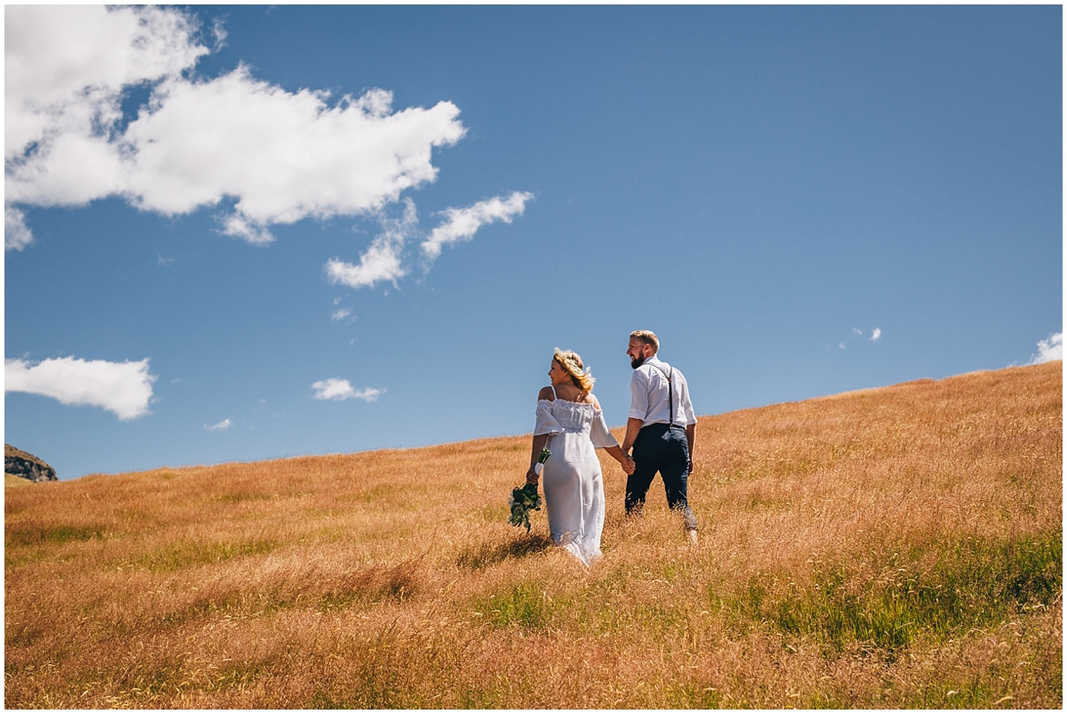 Bride and groom walk across a hillside with sunny blue skies