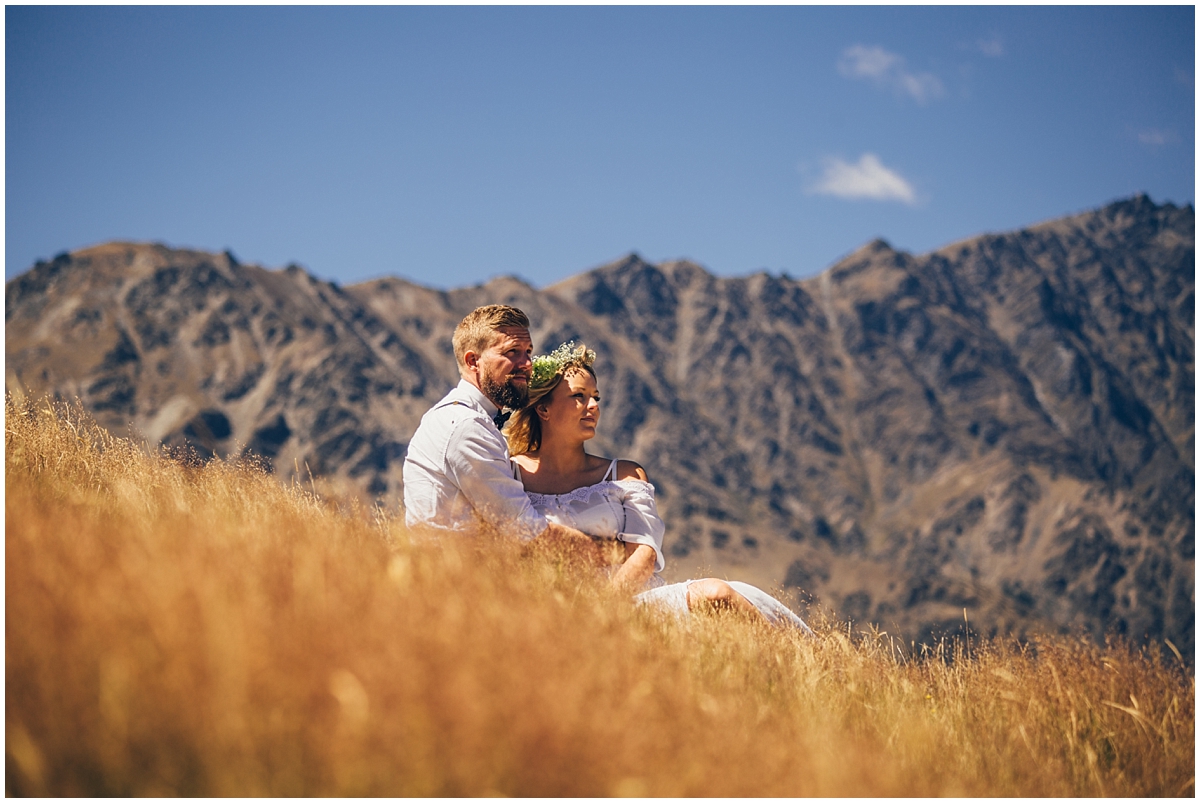 Bride and groom sit on a hillside with mountain backdrop