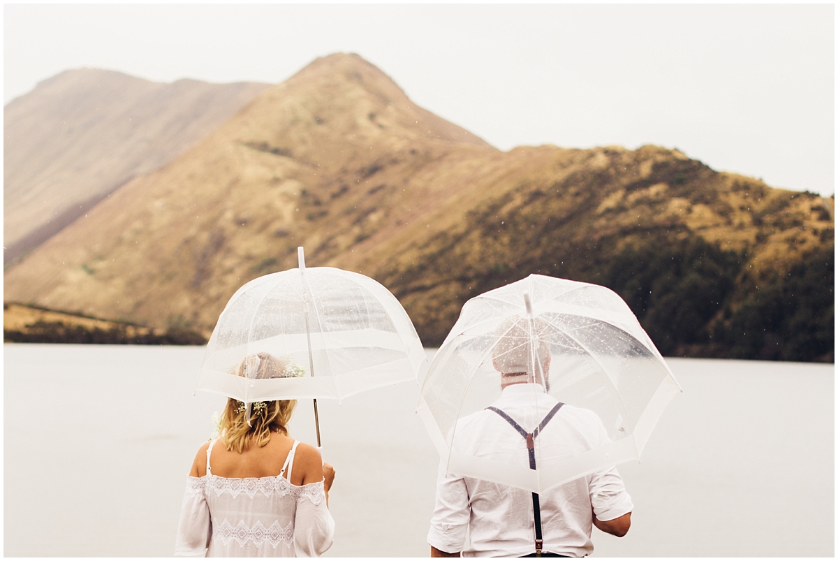 Bride and groom look out over Moke Lake and the mountains under umbrellas