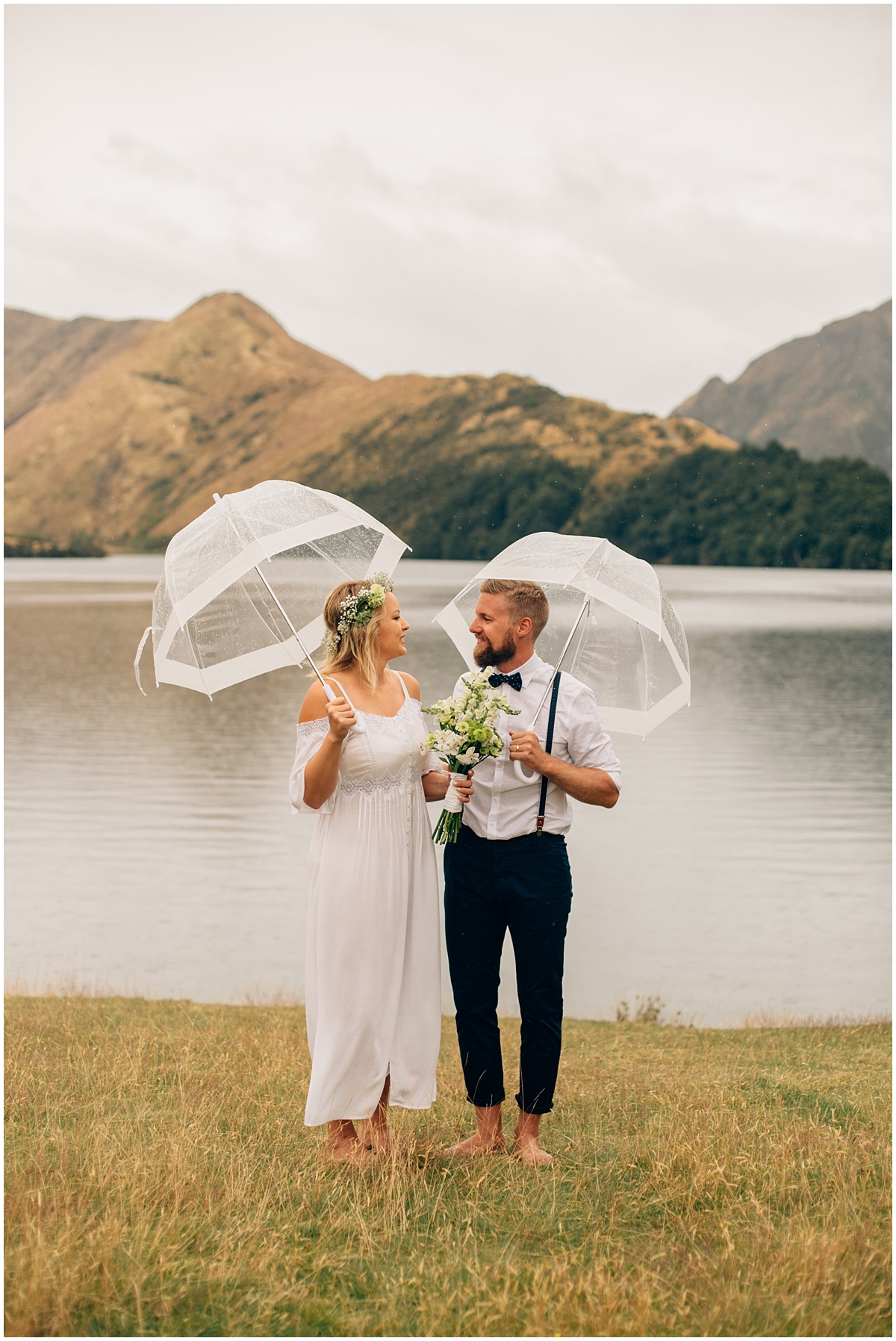 Bride and groom stand at water's edge under umbrellas